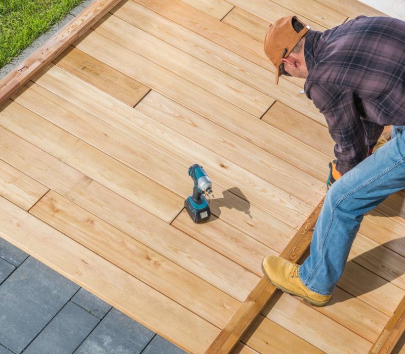 Caucasian Men in His 40s Building Wooden Deck on His Backyard Attaching Wood Board To Each Other Using Cordless Drivers and Screws. Construction Industry Theme.